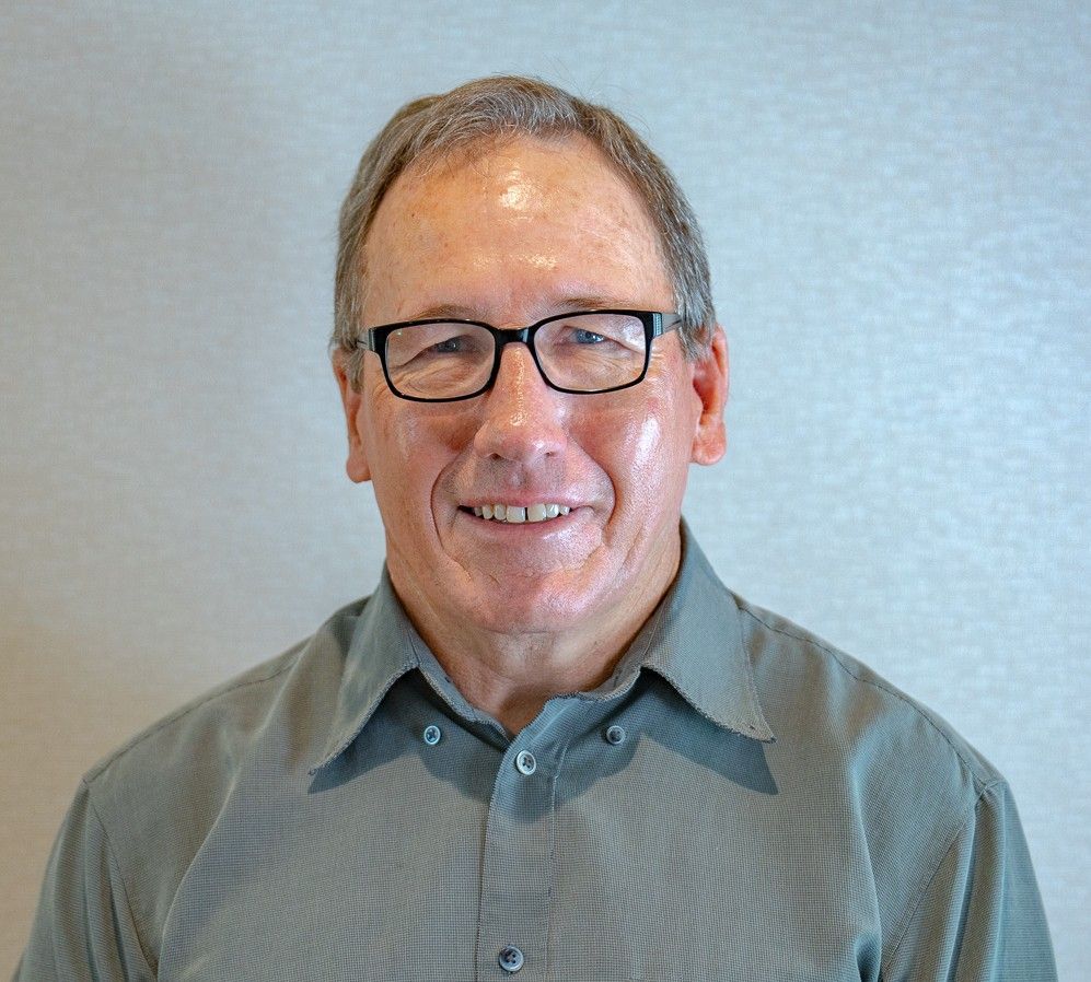 Man with glasses smiles, wearing a gray button-up shirt, against a neutral background.