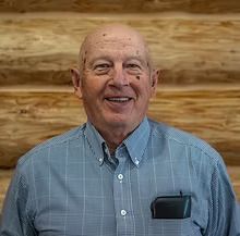 Smiling older man in a blue plaid shirt, posing in front of a wood-paneled wall.
