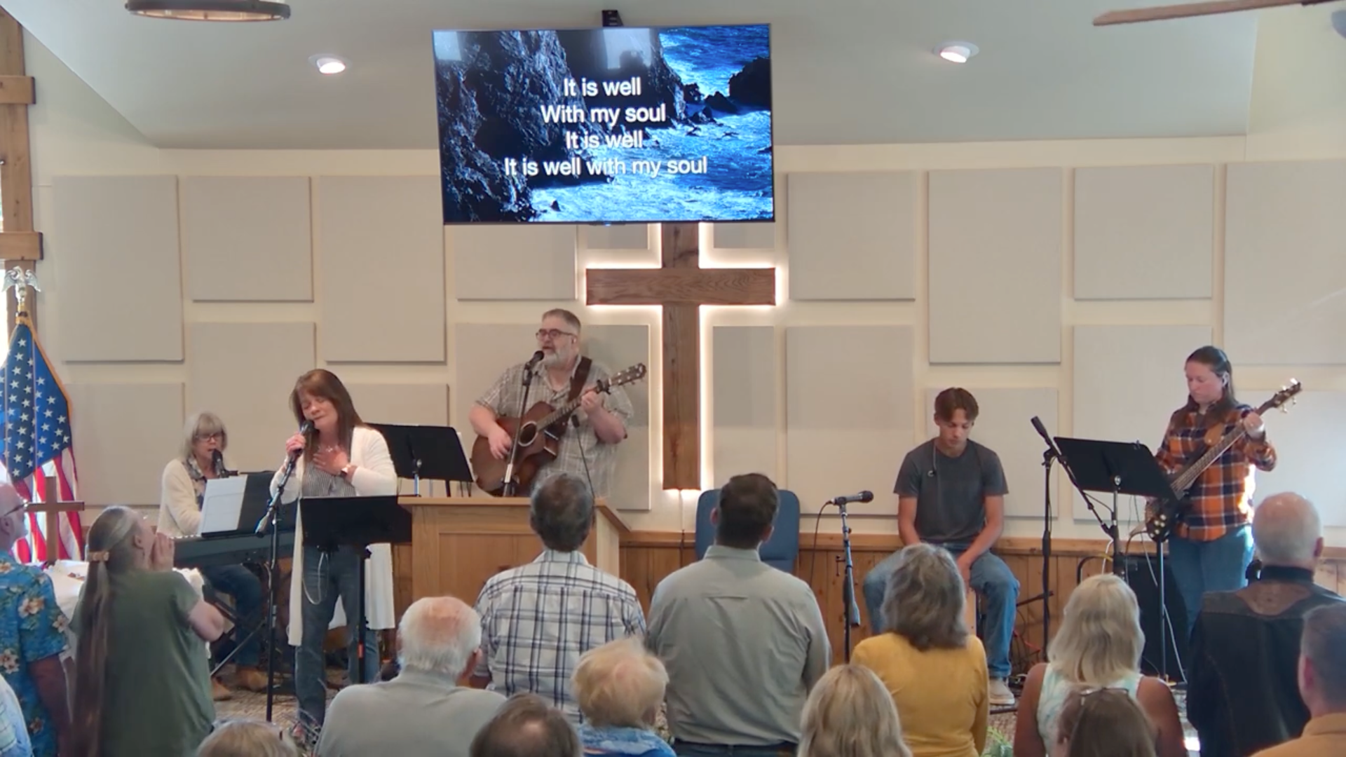 Church service with a band performing; lyrics displayed on a screen; audience in attendance.