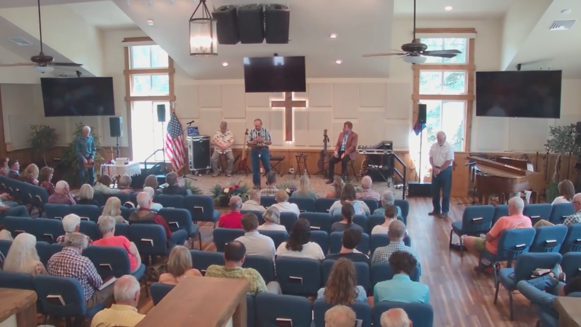 A crowded church service with speakers on a stage and a cross. People are sitting and listening.