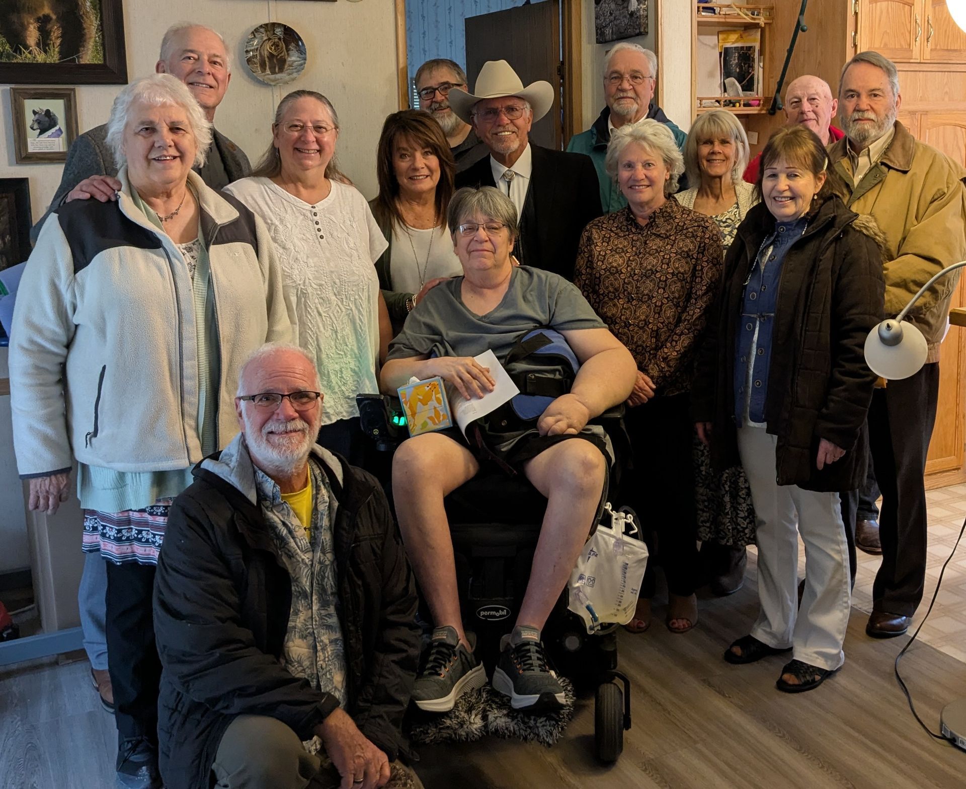 Group photo indoors; people of various ages pose with a man in a wheelchair. Smiling faces.