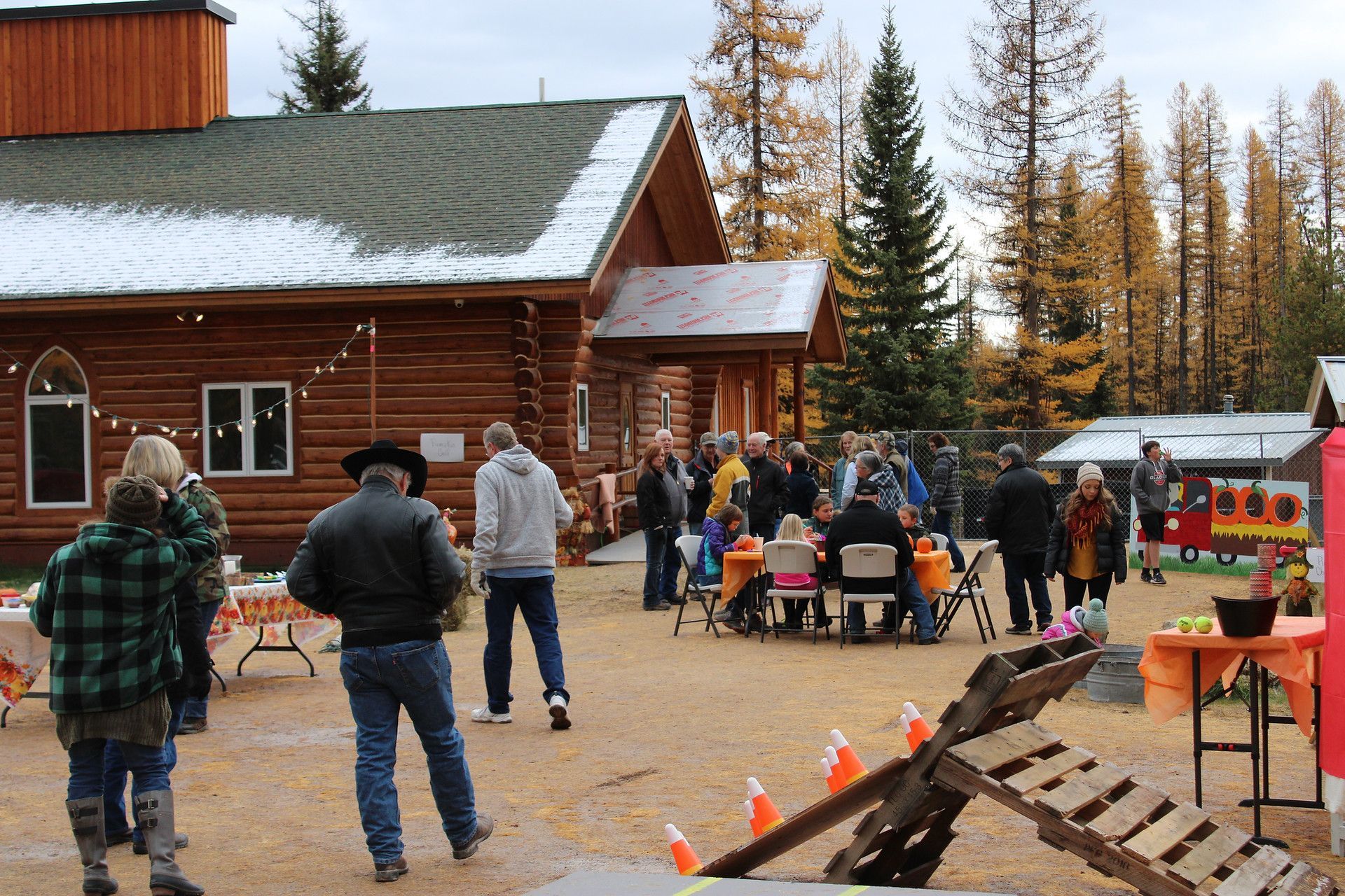 Outdoor gathering near a log cabin. People mingling, some seated. Fall colors, overcast sky.