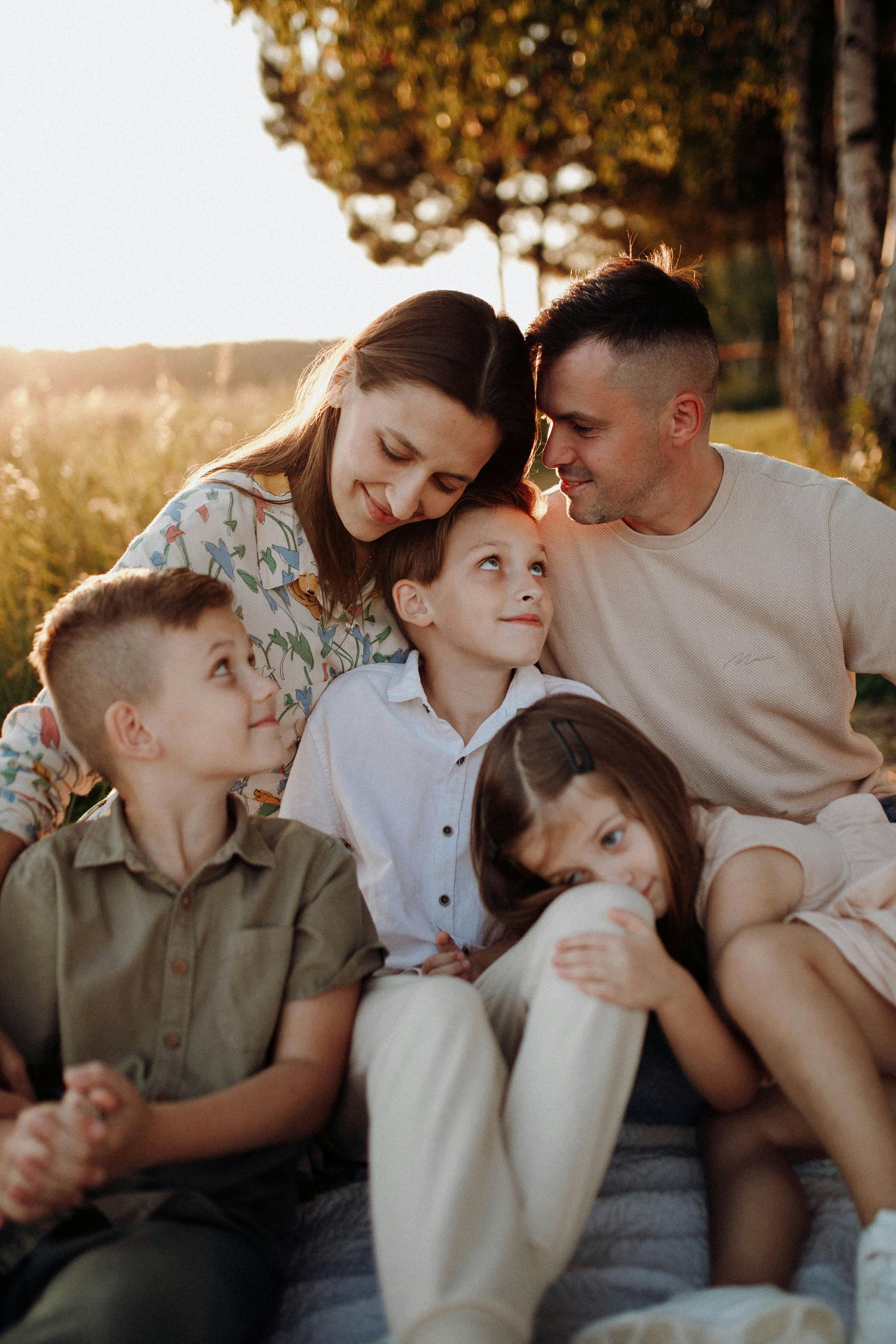 Family of five smiles, cuddled together outdoors; warm sunlight, grassy field.