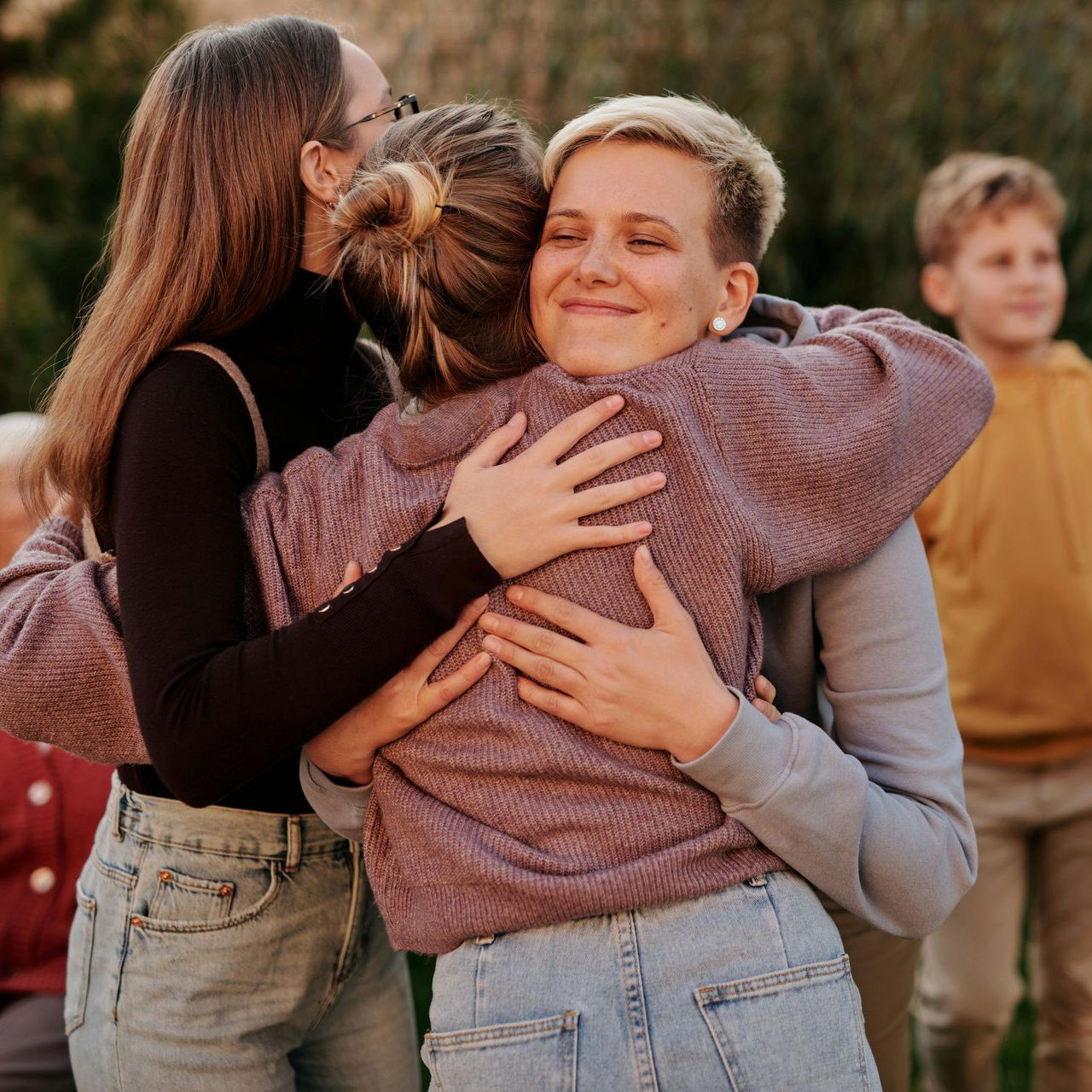 Three women embrace outdoors. One has short blonde hair and smiles. Others wear sweaters, jeans.