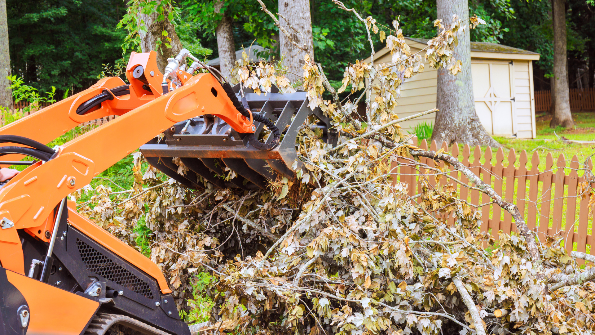 Orange skid steer loader with grapple attachment moving a pile of tree branches.