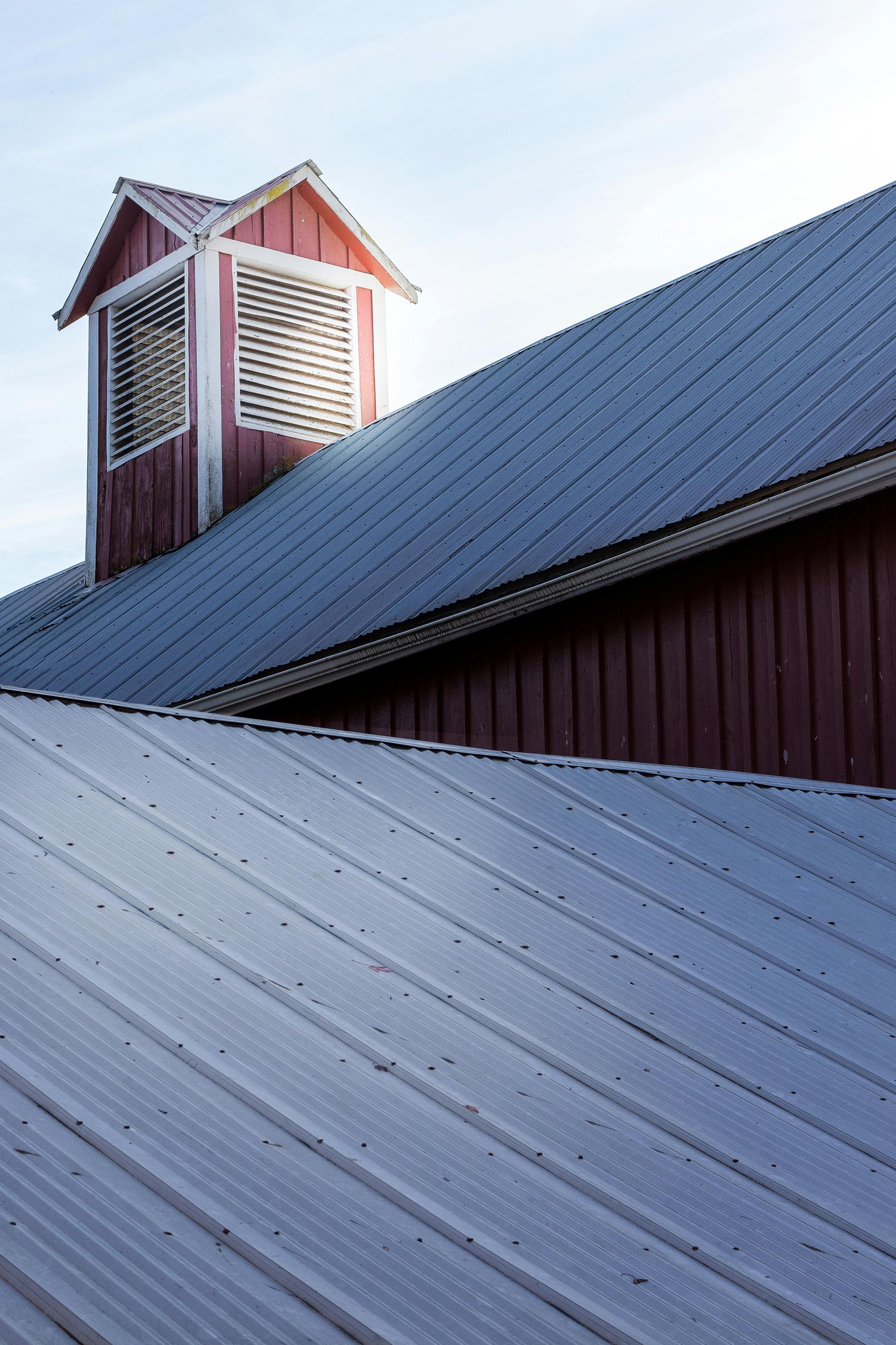 A red barn with a white louvered cupola and a silver metal roof, positioned behind another silver metal roof.
