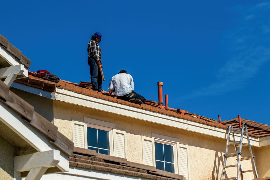Two workers installing tiles on a residential roof under a clear blue sky.