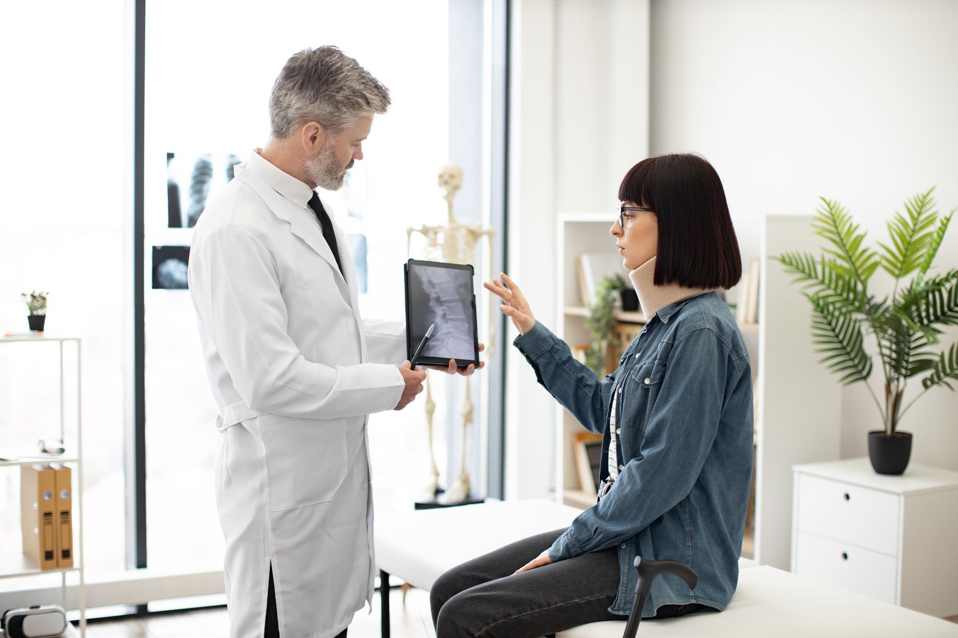 Man in lab coat holding digital tablet while young lady in cervical collar sitting in exam room.