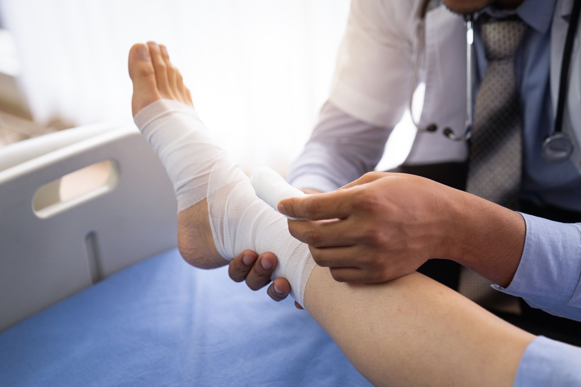 Doctor wrapping a patient's bandaged ankle. Blue sheet and sunlight in the background.