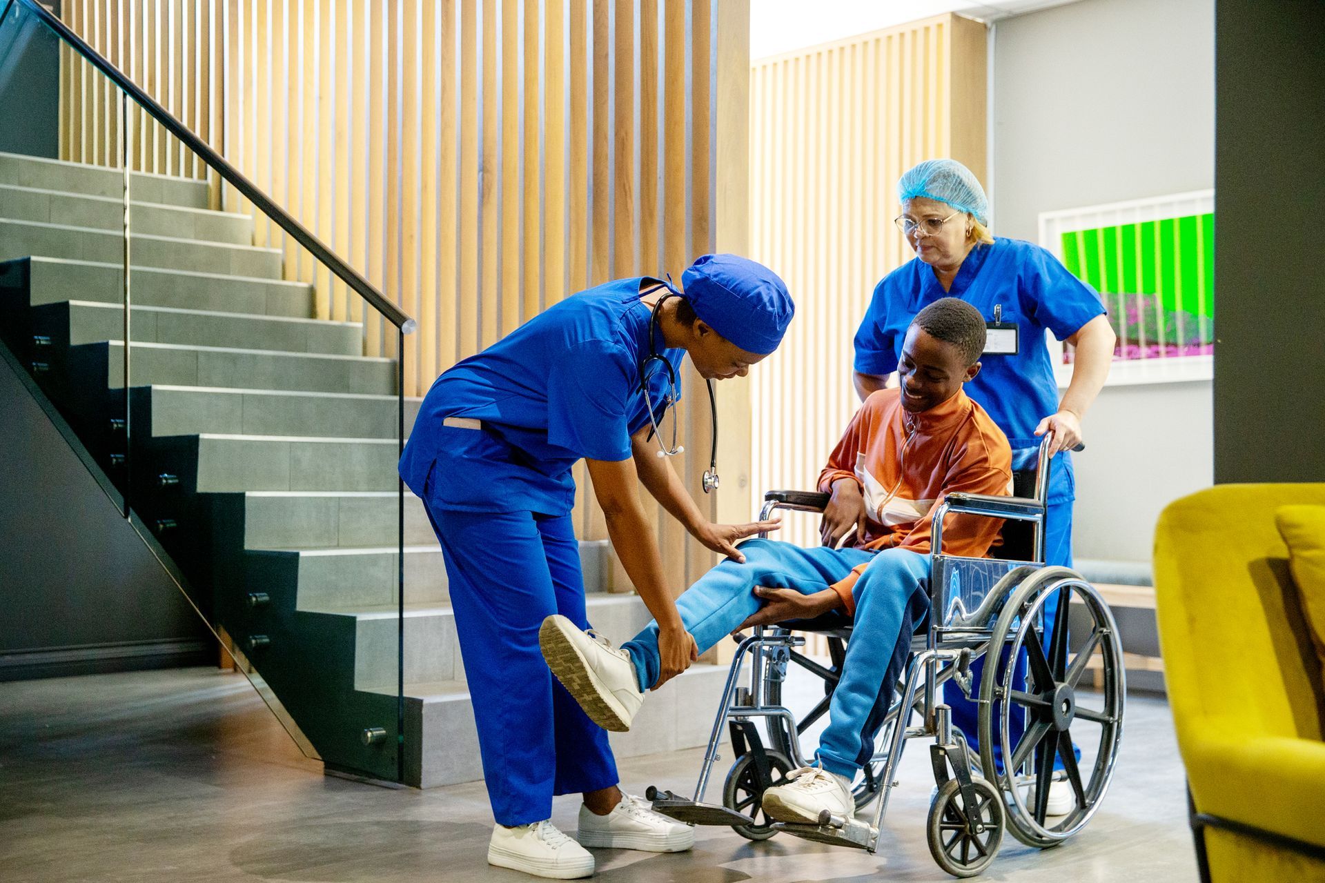 Two nurses helping a person in a wheelchair in a medical setting, near stairs.