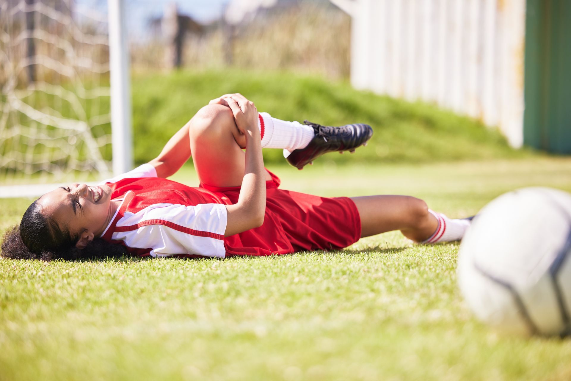Soccer player lying on field, clutching knee in pain. Red uniform, green grass, soccer ball.