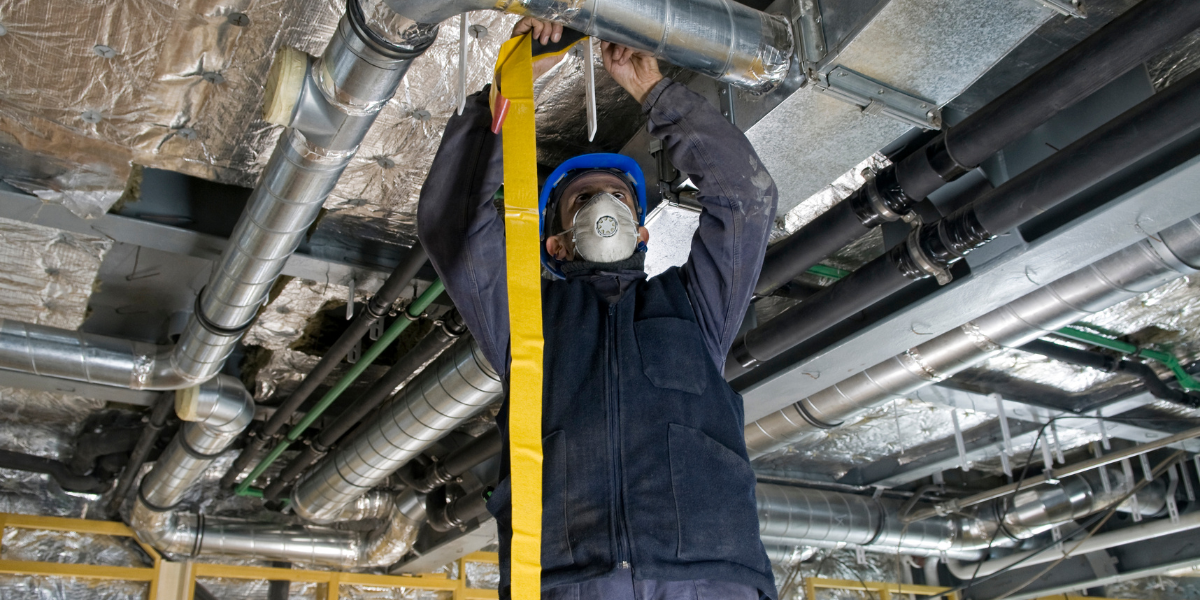 A worker wearing a hard hat and mask working on pipes in an industrial setting.
