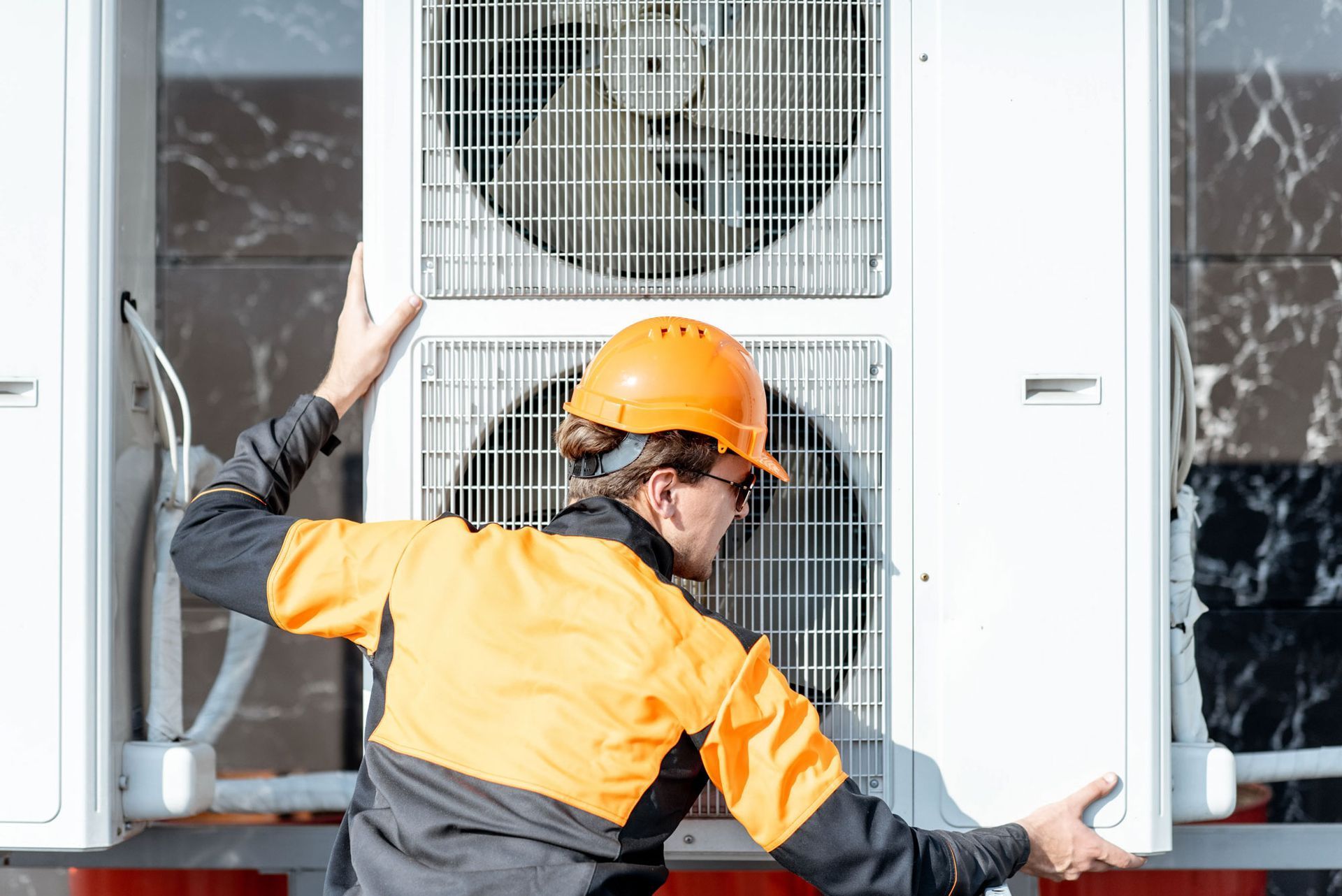 A person in safety gear is inspecting an air conditioning unit outside on a building.