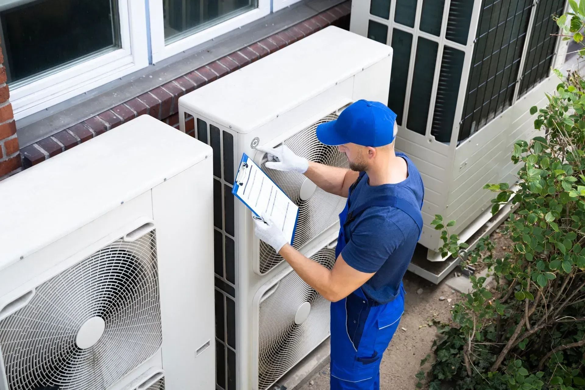 HVAC technician inspecting air conditioning units outside a building, wearing blue uniform and hat.