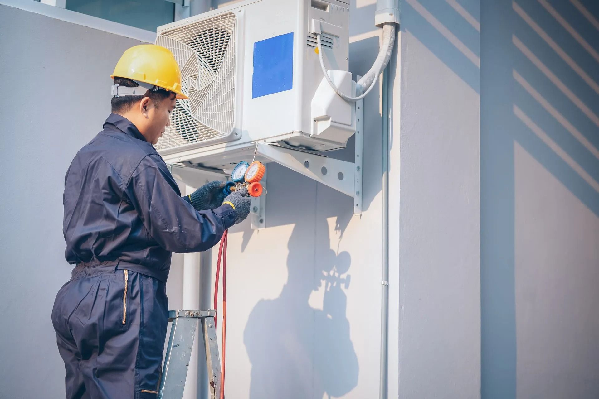 HVAC technician in blue jumpsuit and yellow hard hat using a multimeter to service an outdoor air conditioning unit.