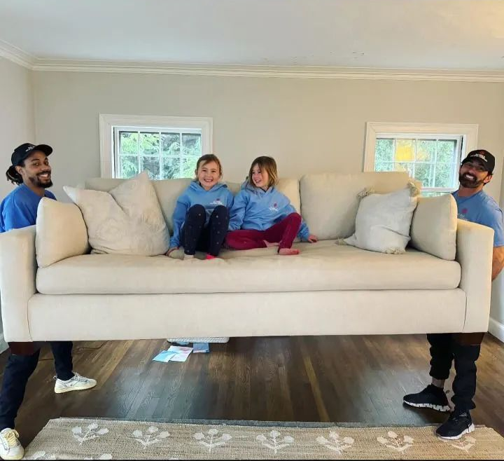 Four men carrying a beige sofa with two children sitting on it. Indoors with windows and hardwood floors.