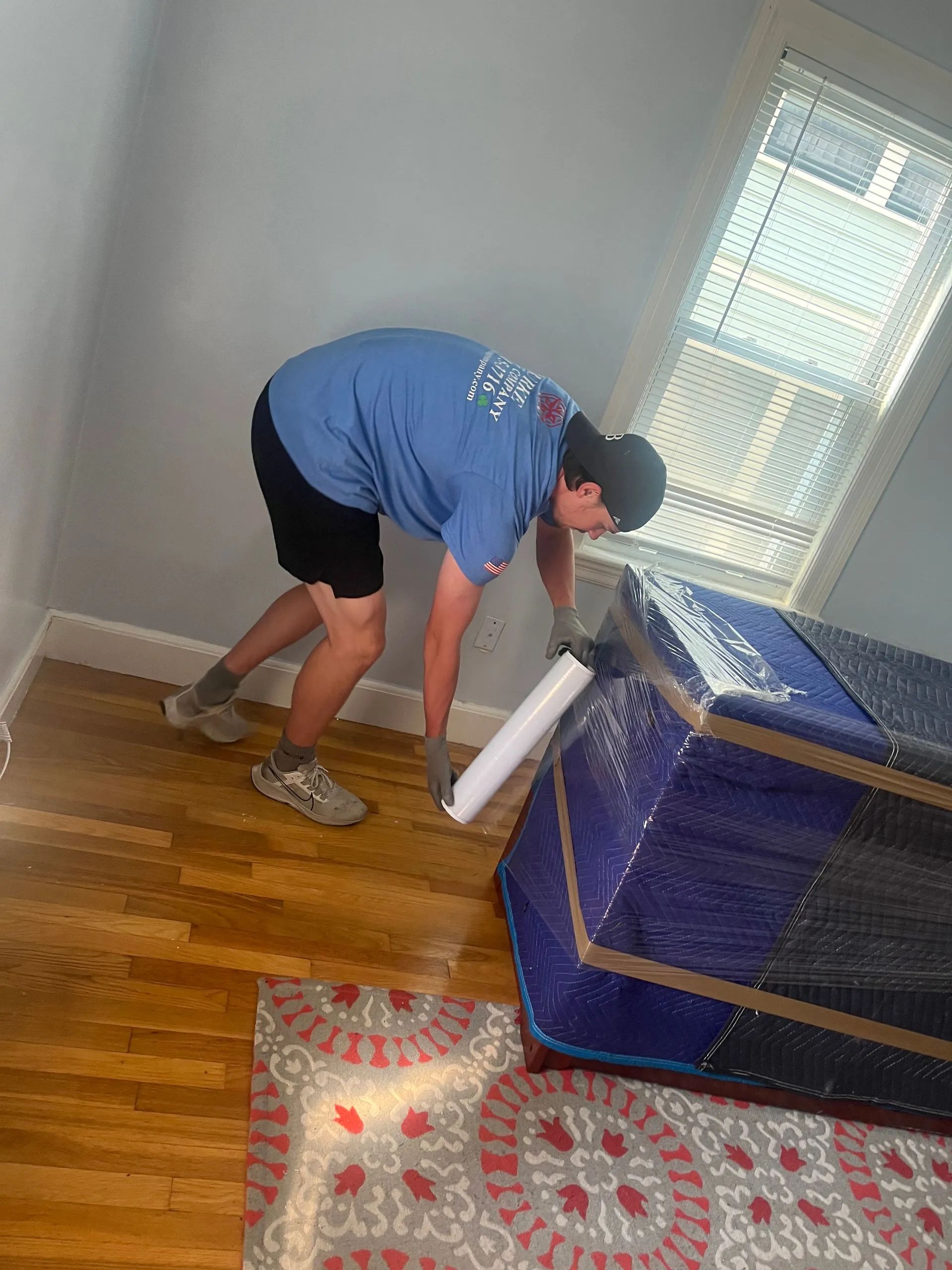 A person in a blue shirt and black shorts wraps a piece of furniture in protective blue moving plastic in a room.