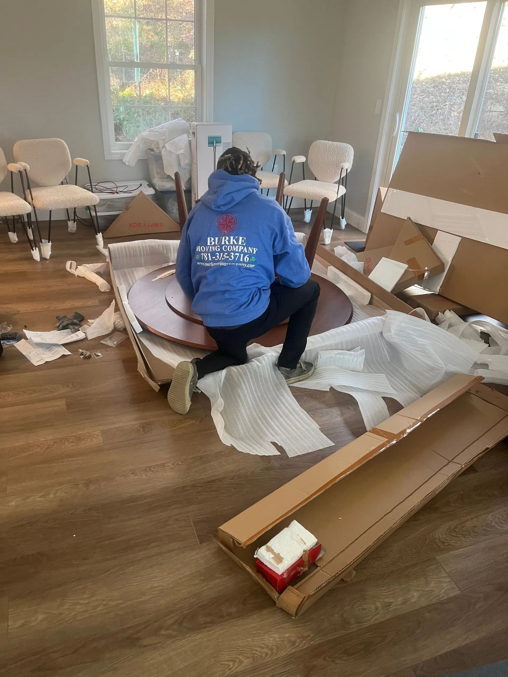 Person kneeling, assembling a round dining table in a room with cardboard boxes and chairs.