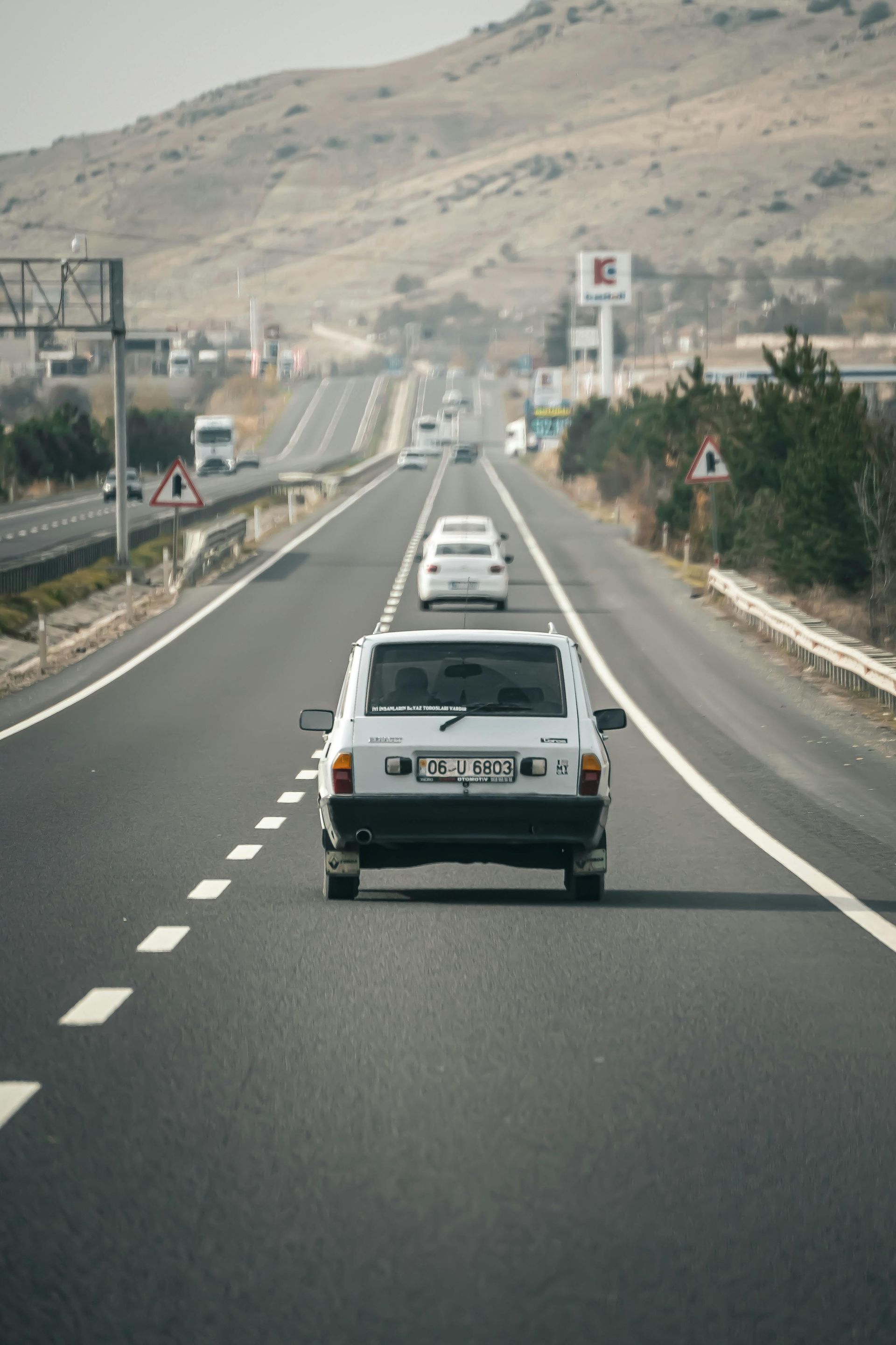 White car driving on a highway, other vehicles visible in the distance, hills in the background.