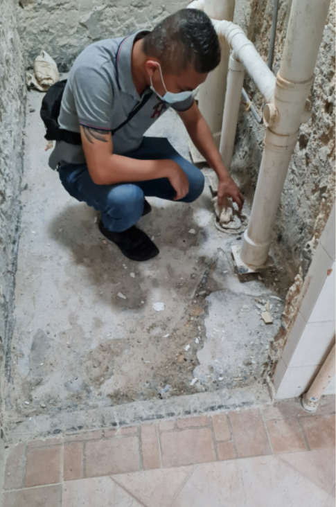 A man wearing a mask is kneeling on the floor looking at a pipe.