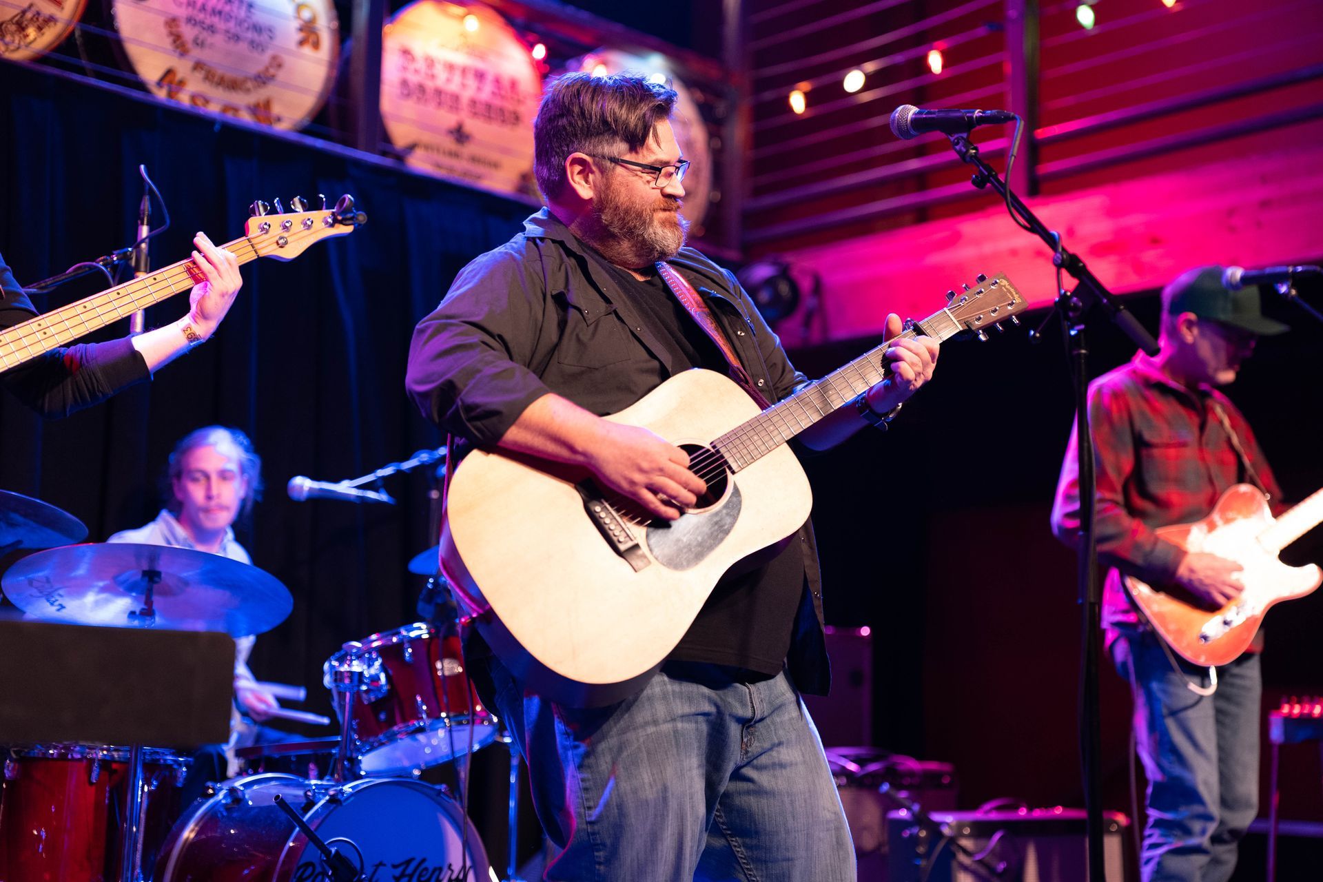 Band performing live on stage; guitarist center, drums, bass, and additional guitarists. Stage lit with purple and red hues.