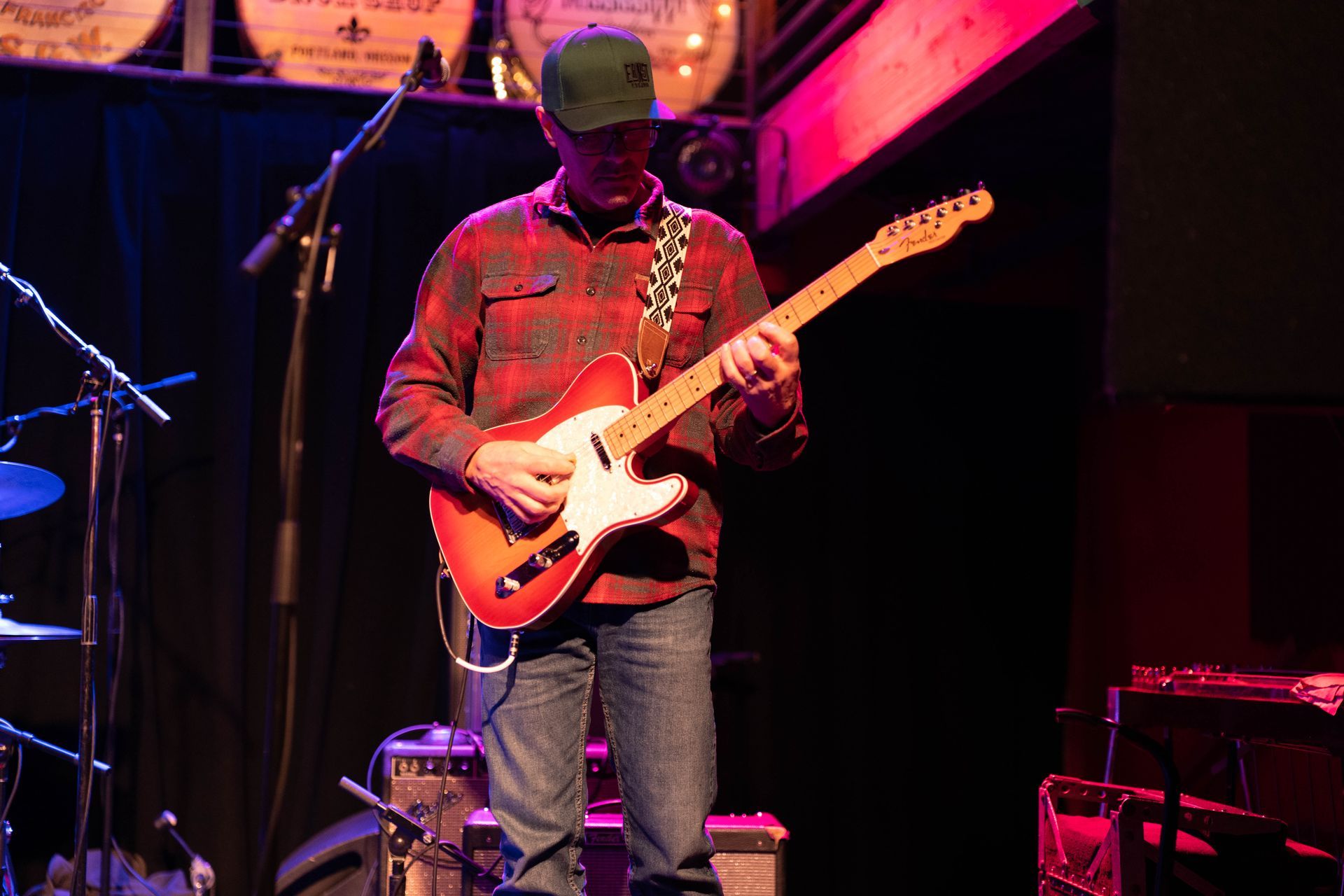 Man playing red and white electric guitar on stage, wearing plaid shirt and jeans.