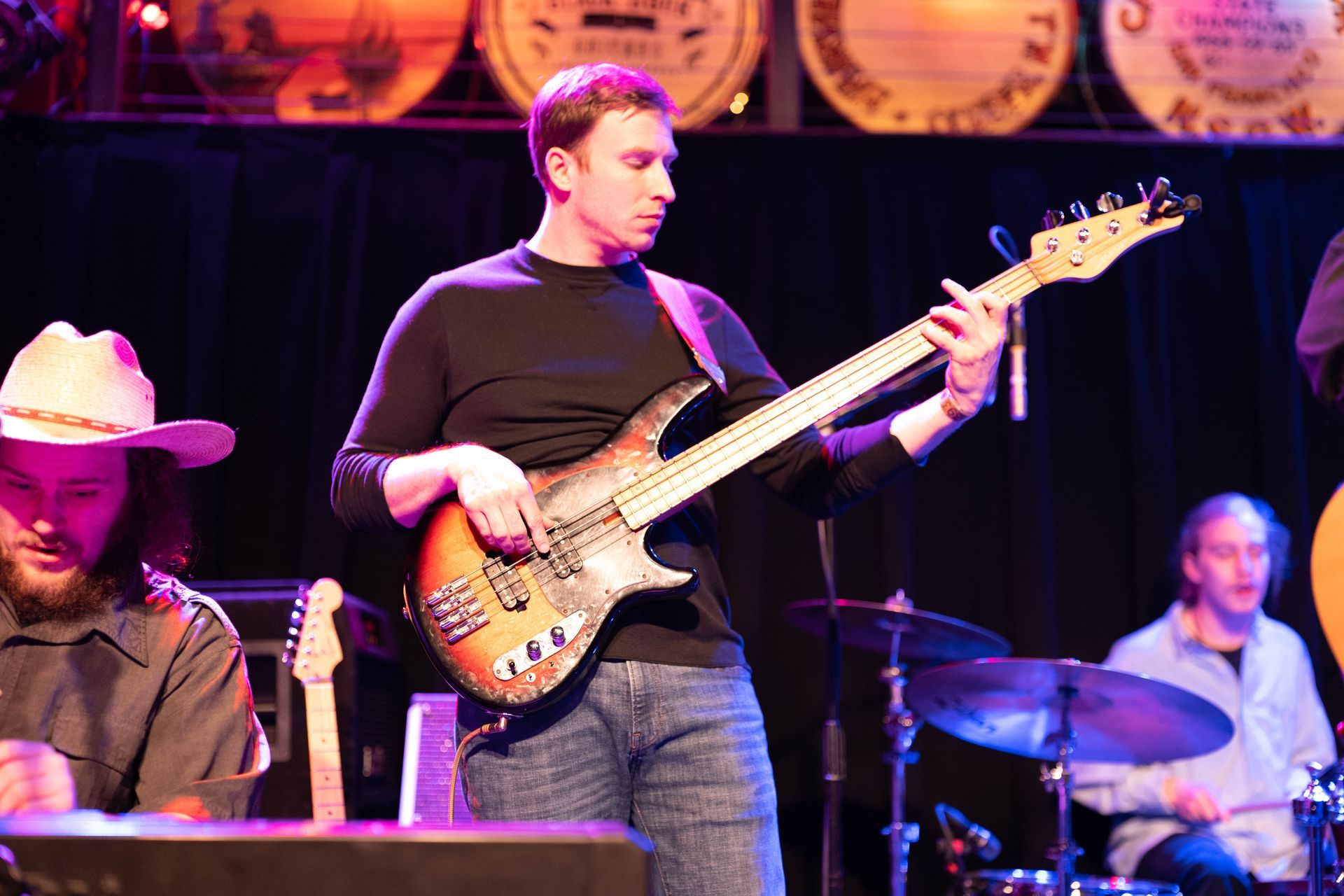 Man playing bass guitar onstage, bandmates visible. Dark setting with barrels as backdrop.