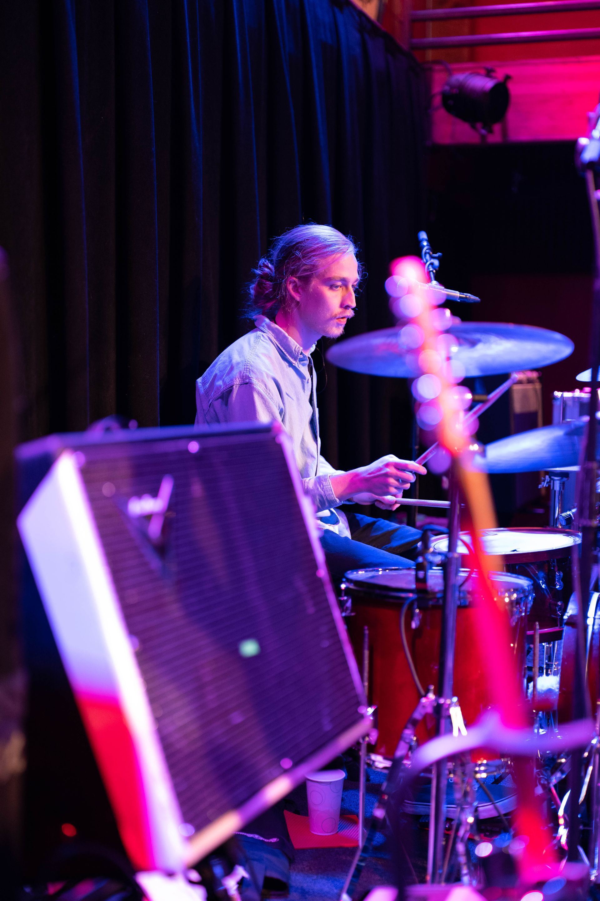 Drummer with long hair playing on stage, illuminated by purple light.