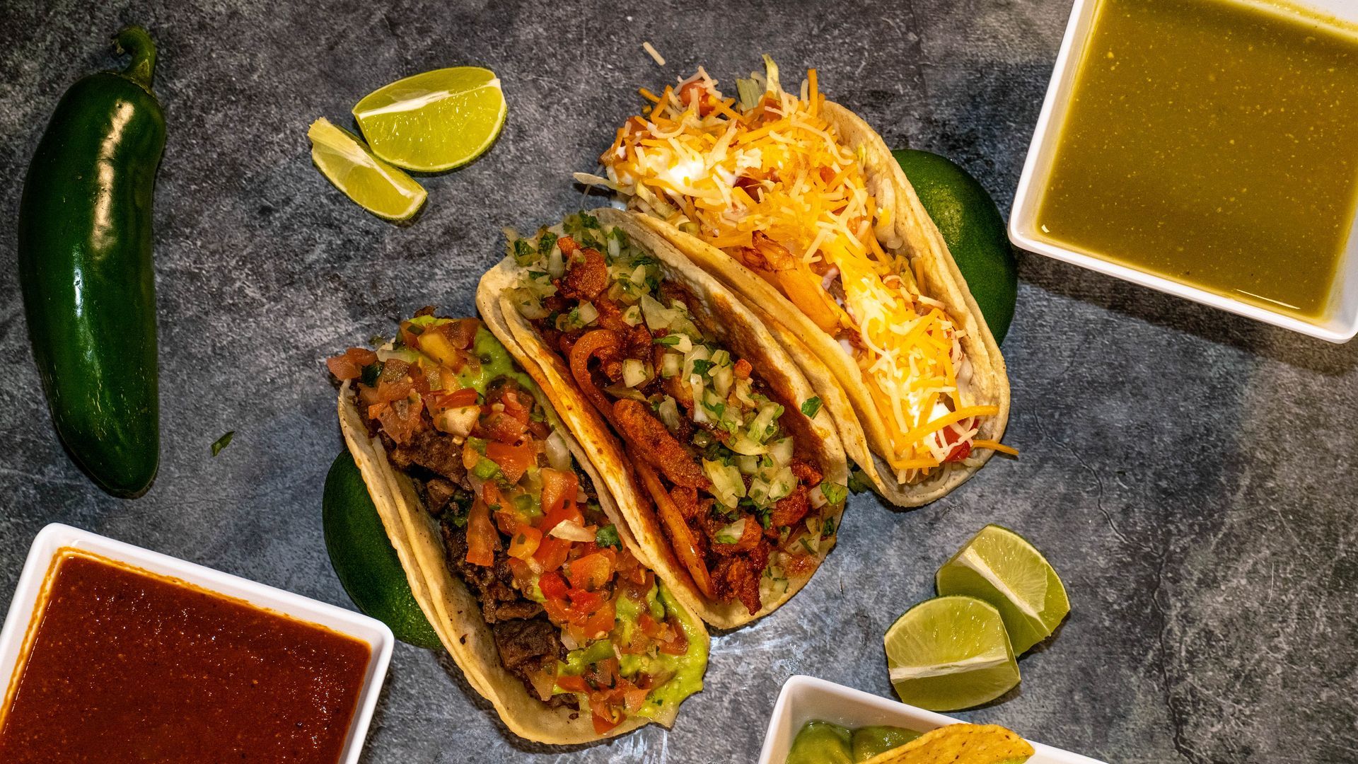Three tacos with various fillings, surrounded by lime wedges, sauces, and a jalapeño on a textured surface.