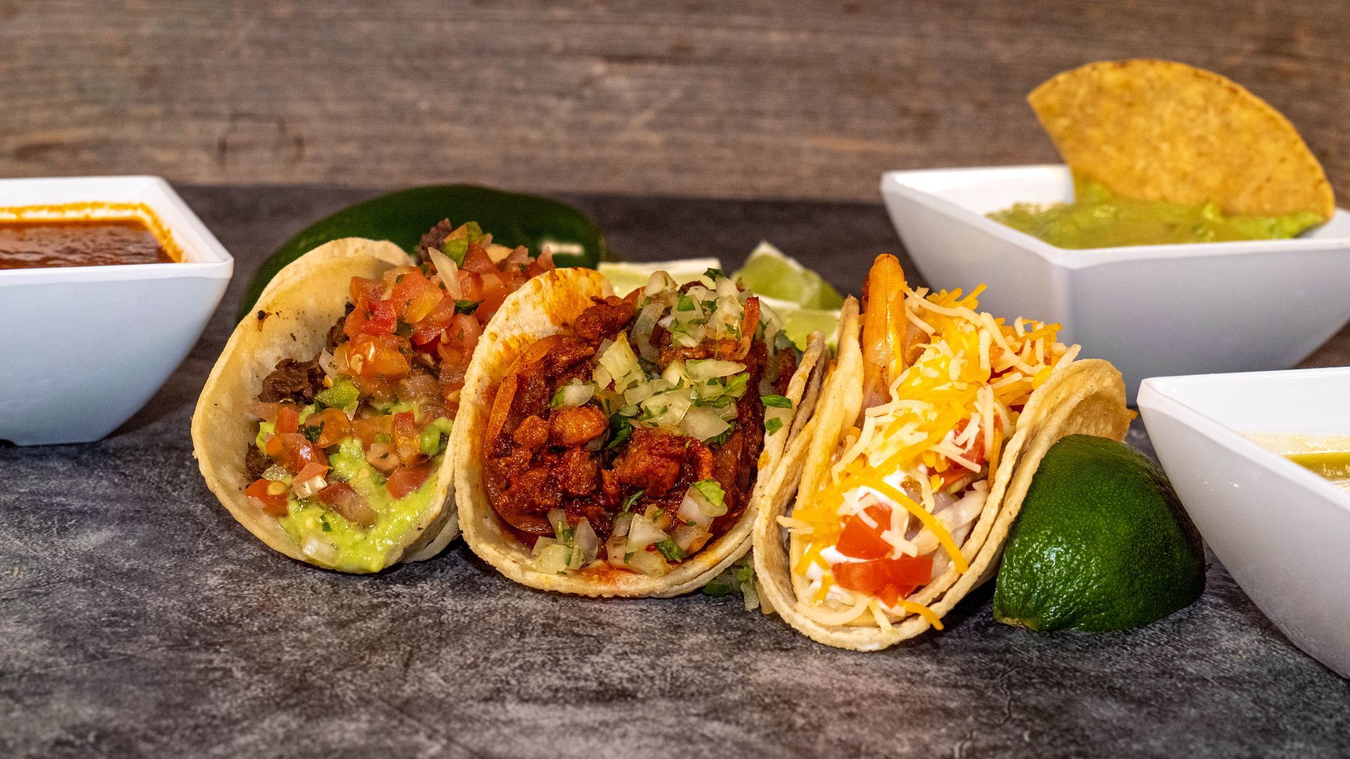 Tacos with various fillings, guacamole, salsa, and lime on a gray surface.
