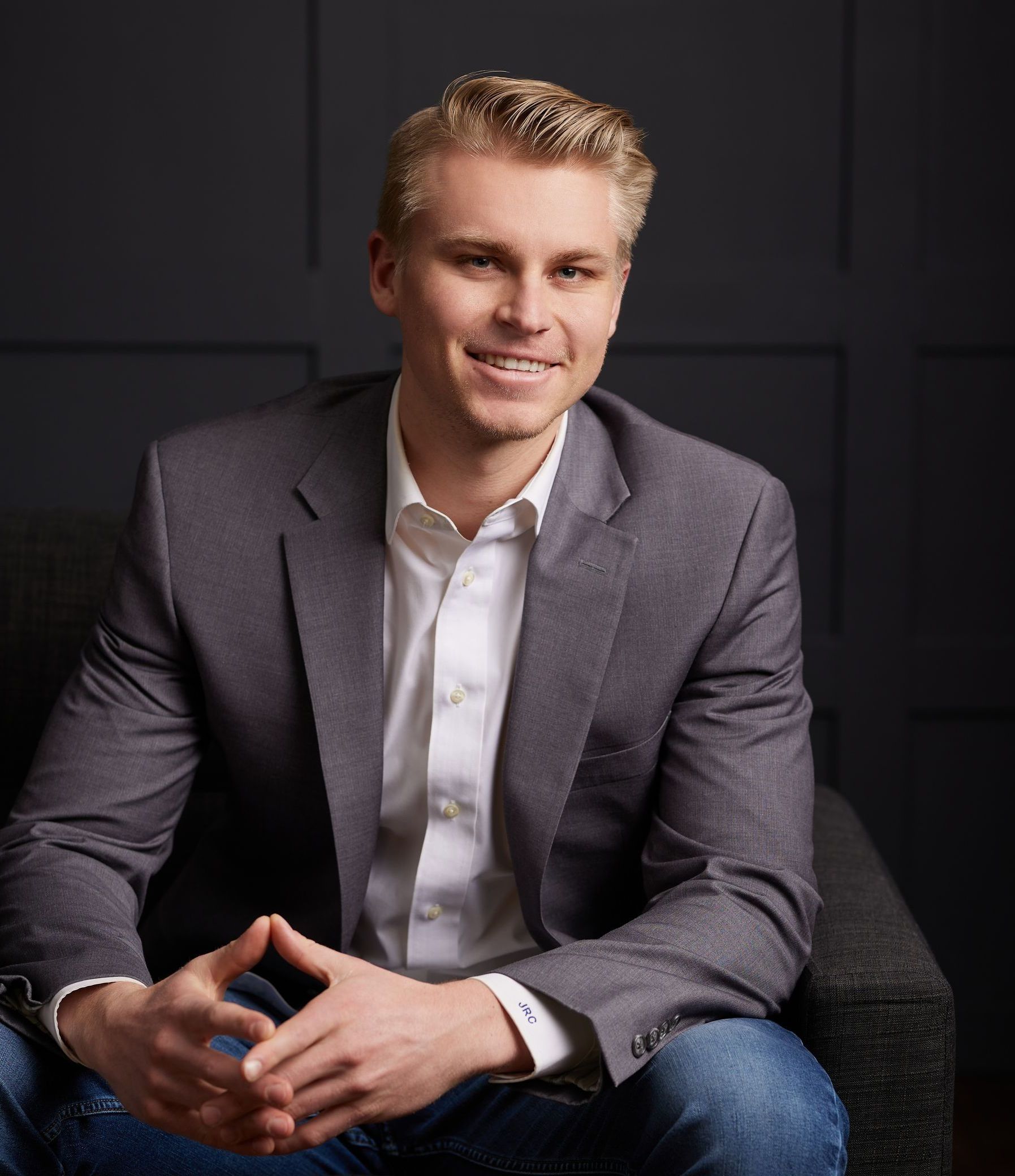 A man in a suit and white shirt is sitting in a chair with his hands folded.