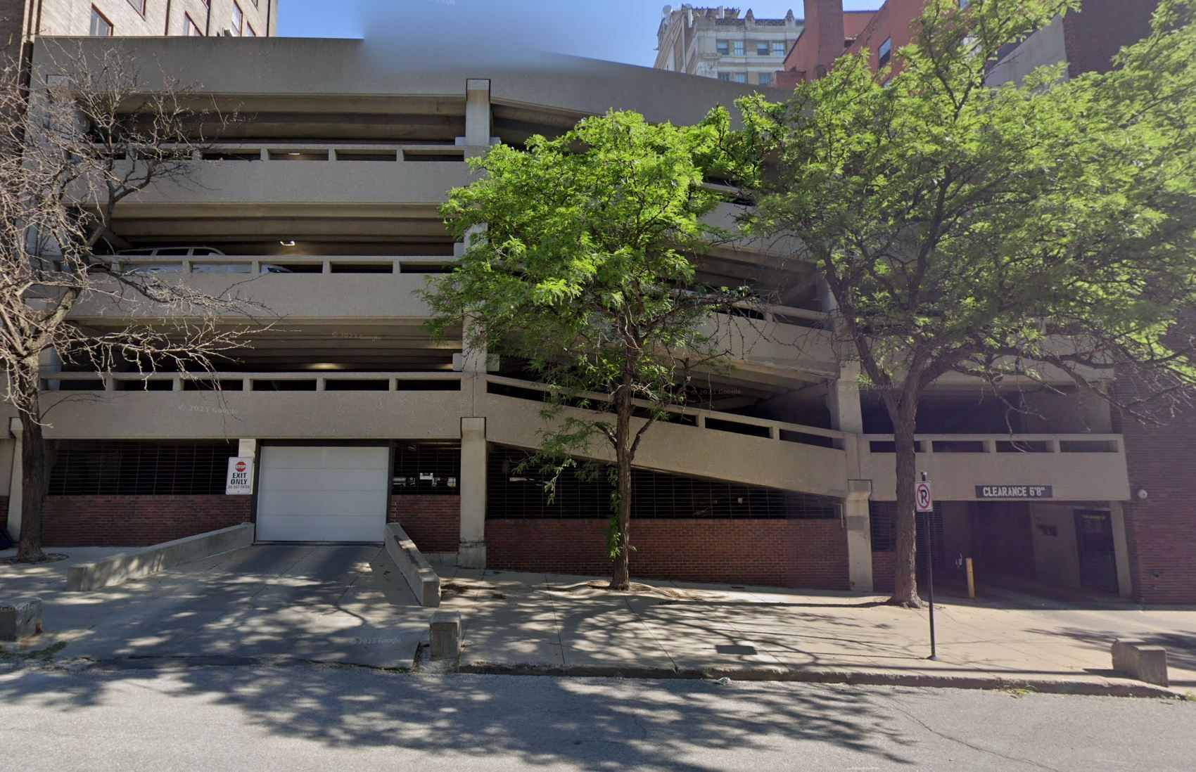 Multi-story parking garage with sloping ramps. Entrance is on the left. Trees in the middle, and building on the right.