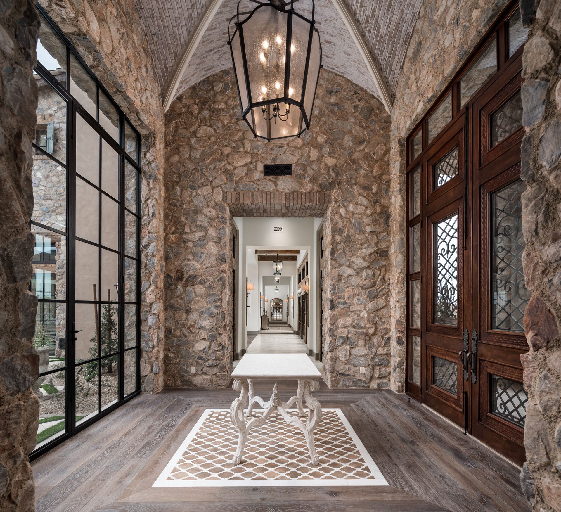 Stone-Walled Entrance with Large Windows: An impressive entrance hallway with stone walls, a vaulted ceiling, and large black-framed windows offering a view of the mountains. The space features decorative iron doors, a central bench, and a large lantern-style chandelier. The floor includes a patterned tile design.