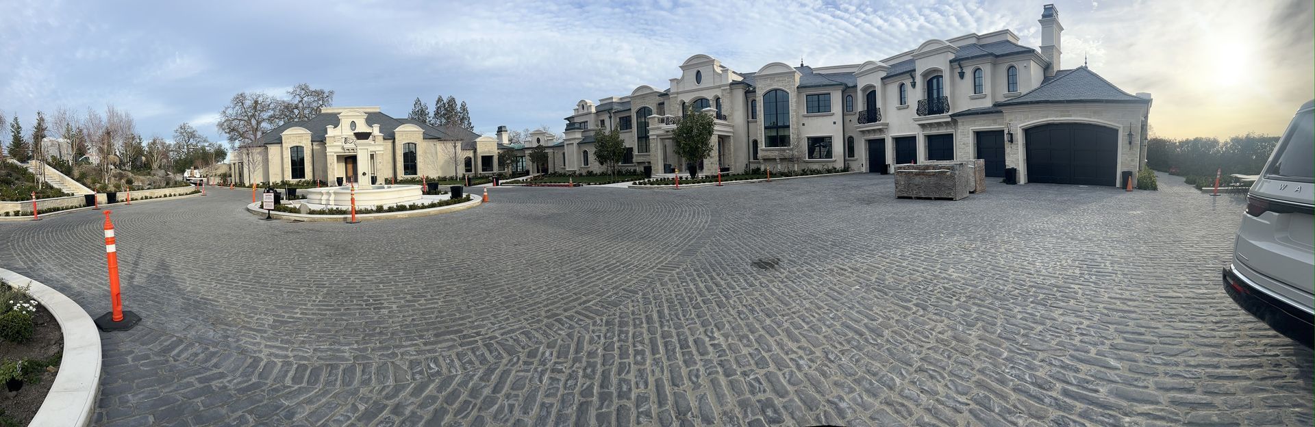 Large, light-colored mansion with a circular driveway, surrounded by greenery and a view of rolling hills.