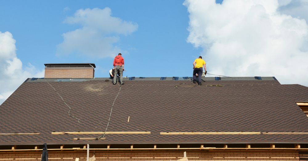 Two roofers on a brown shingled roof under a blue sky, installing roofing material.