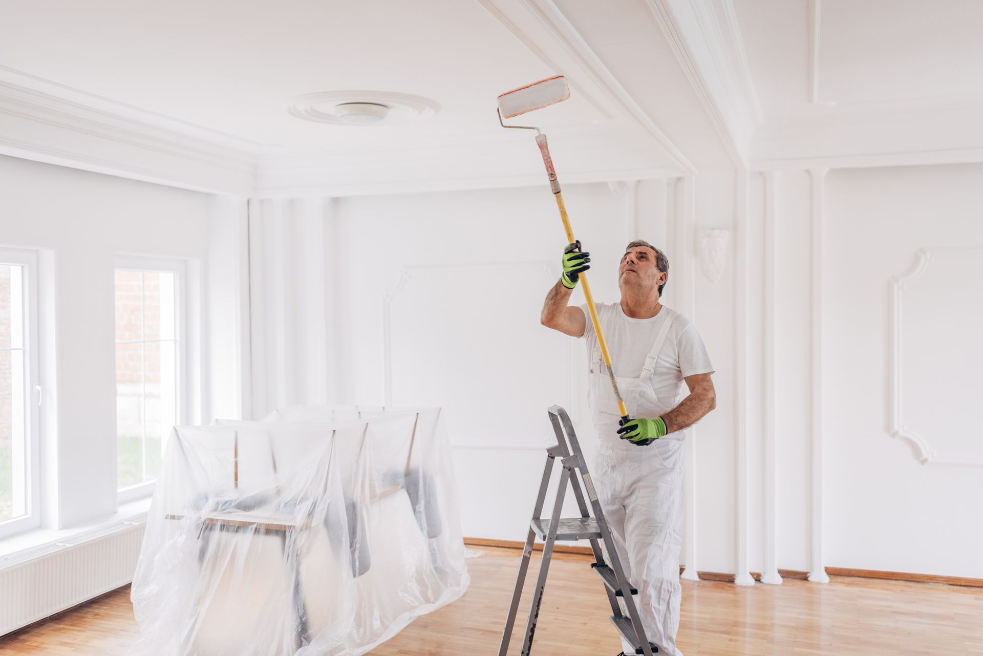 A man is standing on a ladder painting the ceiling of a room.
