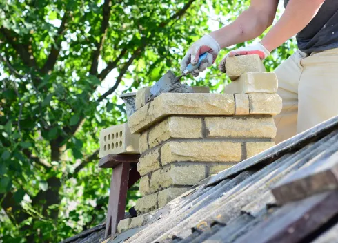 A man is laying bricks on top of a chimney on a roof.