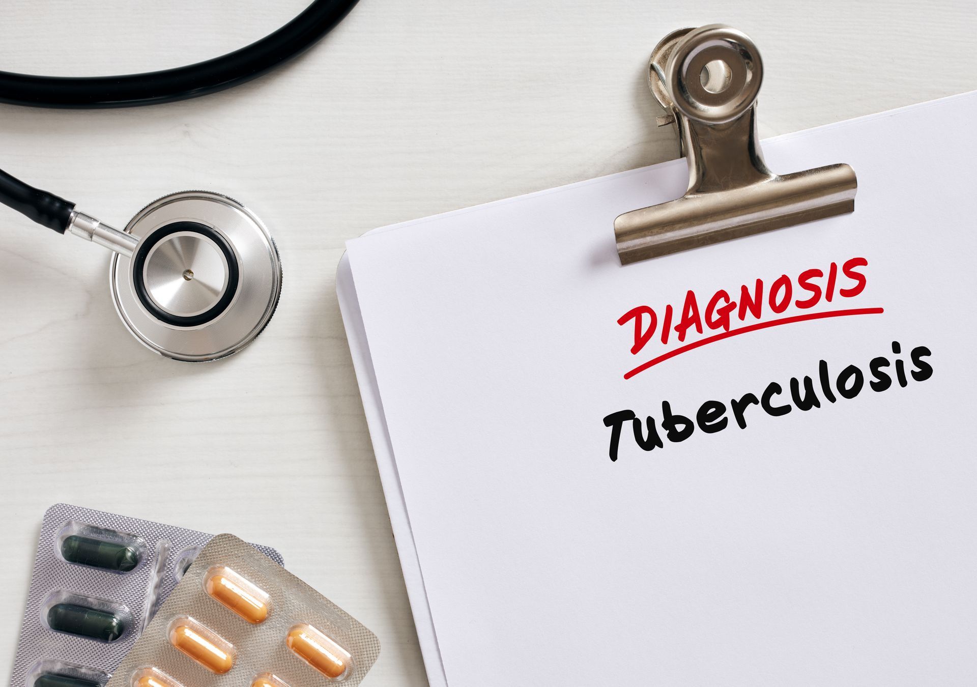 A stethoscope and blister packs of pills lie on a desk next to a clipboard showing the diagnosis: Tuberculosis.
