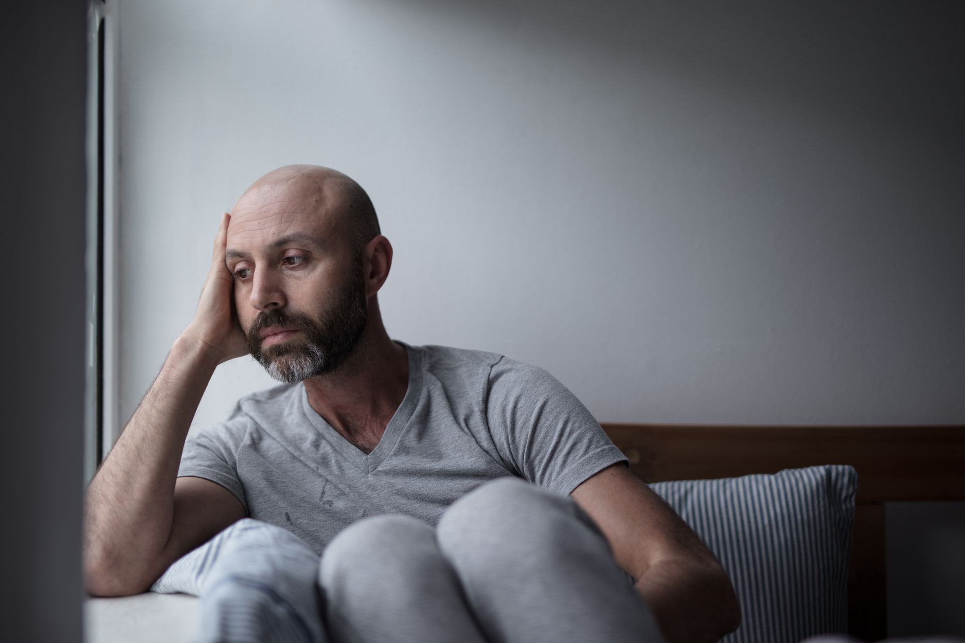 Man with beard, resting hand on head, looking down with a sad expression, seated near a window.
