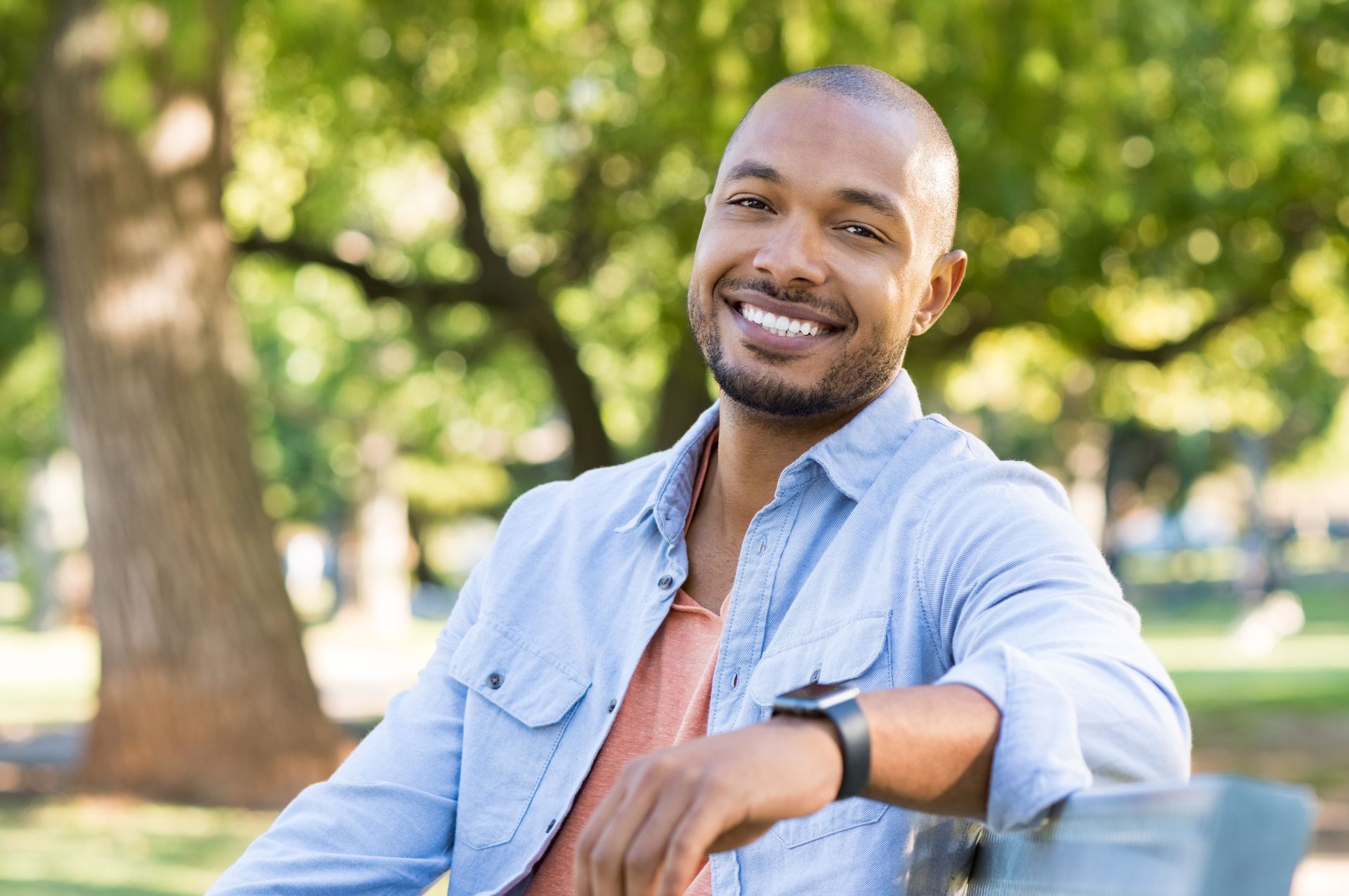 Smiling person in light blue shirt sits on a park bench, sunny outdoors.