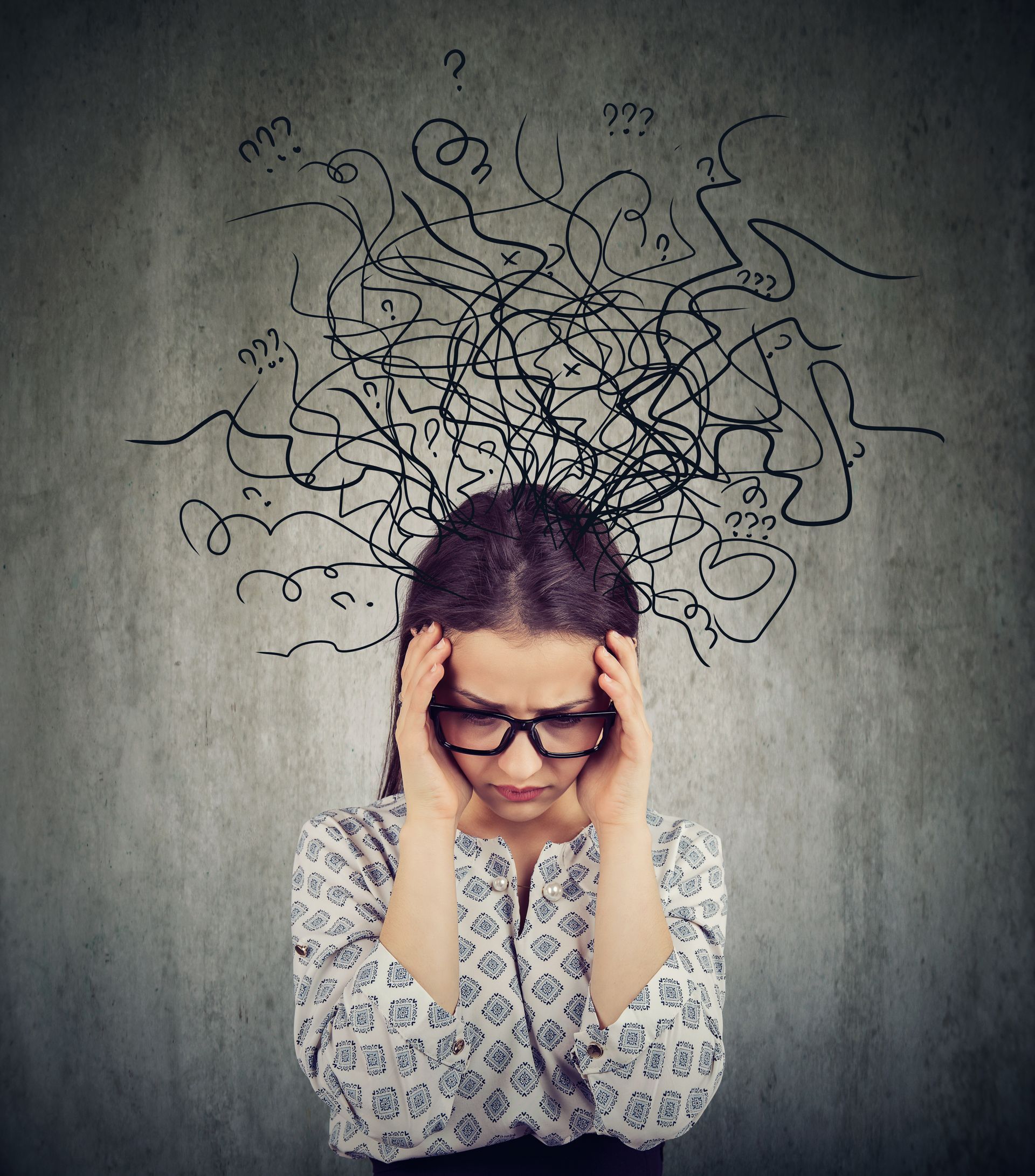 Woman with glasses, hands on head, looking down with a tangled scribble illustration above her head, against a grey background.