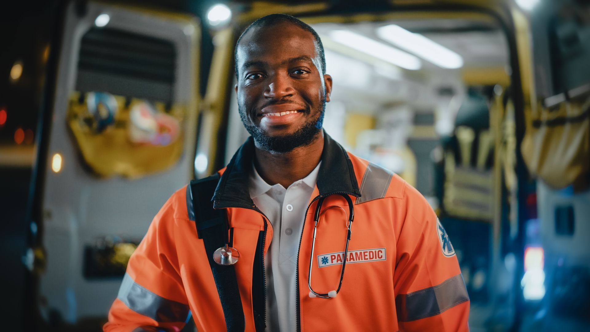Smiling person in orange medical jacket, stethoscope, standing in front of an ambulance with open doors, at night.