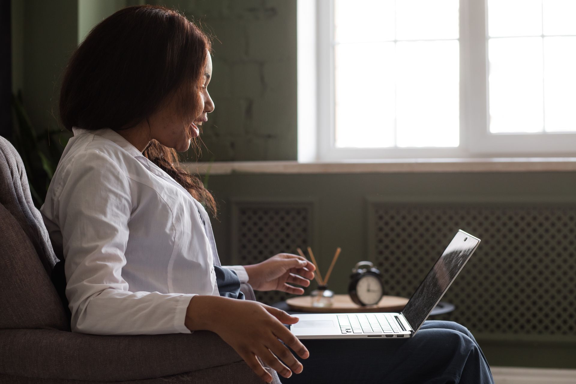 Woman sits in armchair, using laptop near a window.