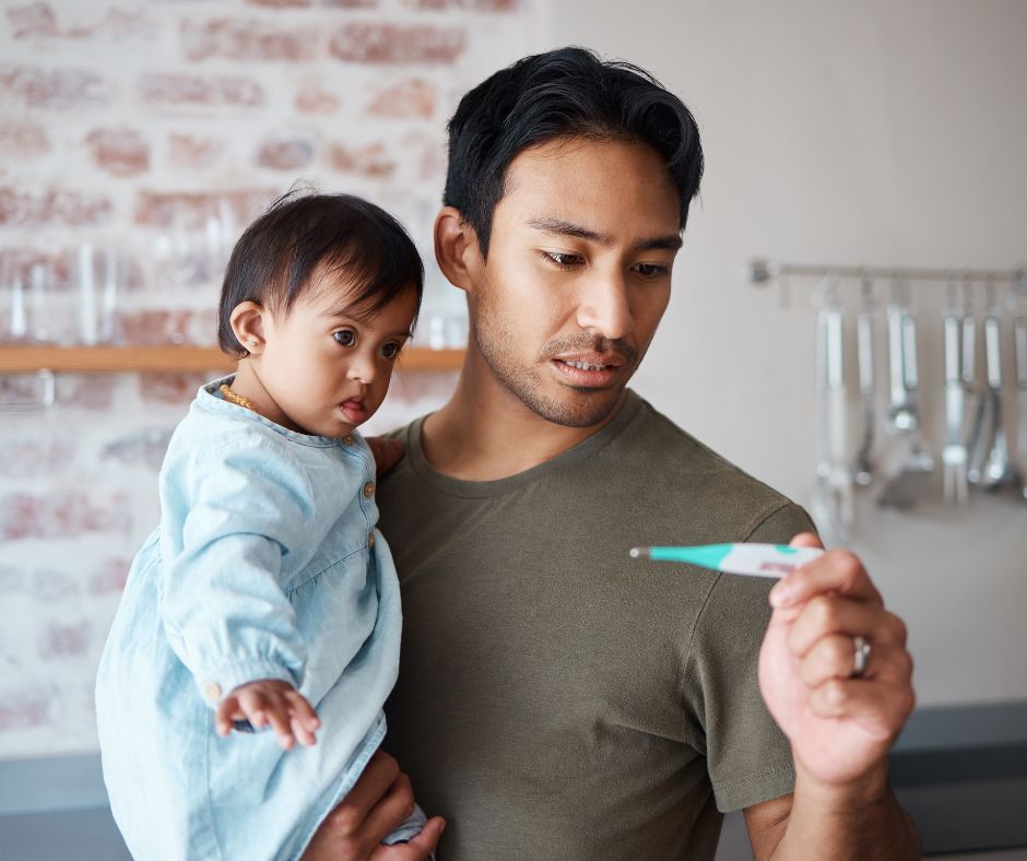 Man holding a baby, looking at a thermometer. Kitchen setting.