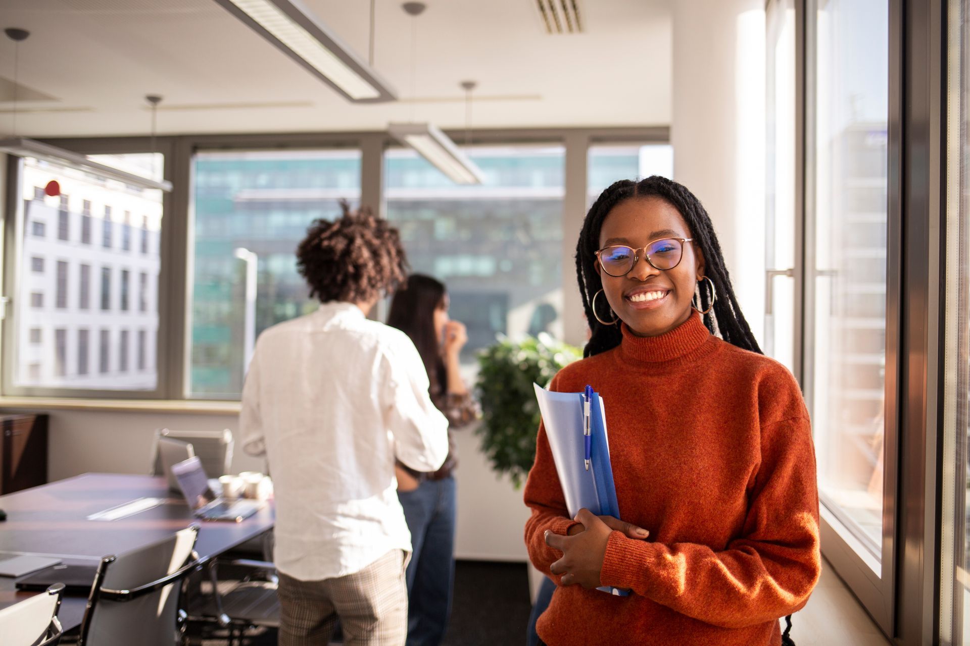 A smiling woman in an orange sweater holds a folder in a bright office while colleagues work in the background.