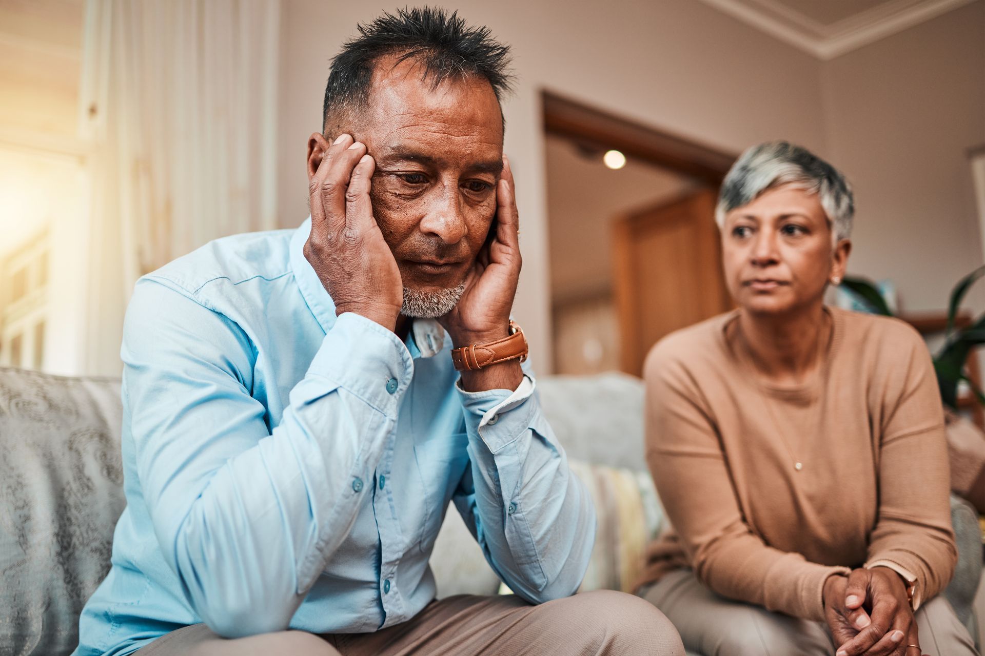 Man with head in hands looks distressed, sitting beside woman looking on; indoors, on a couch.