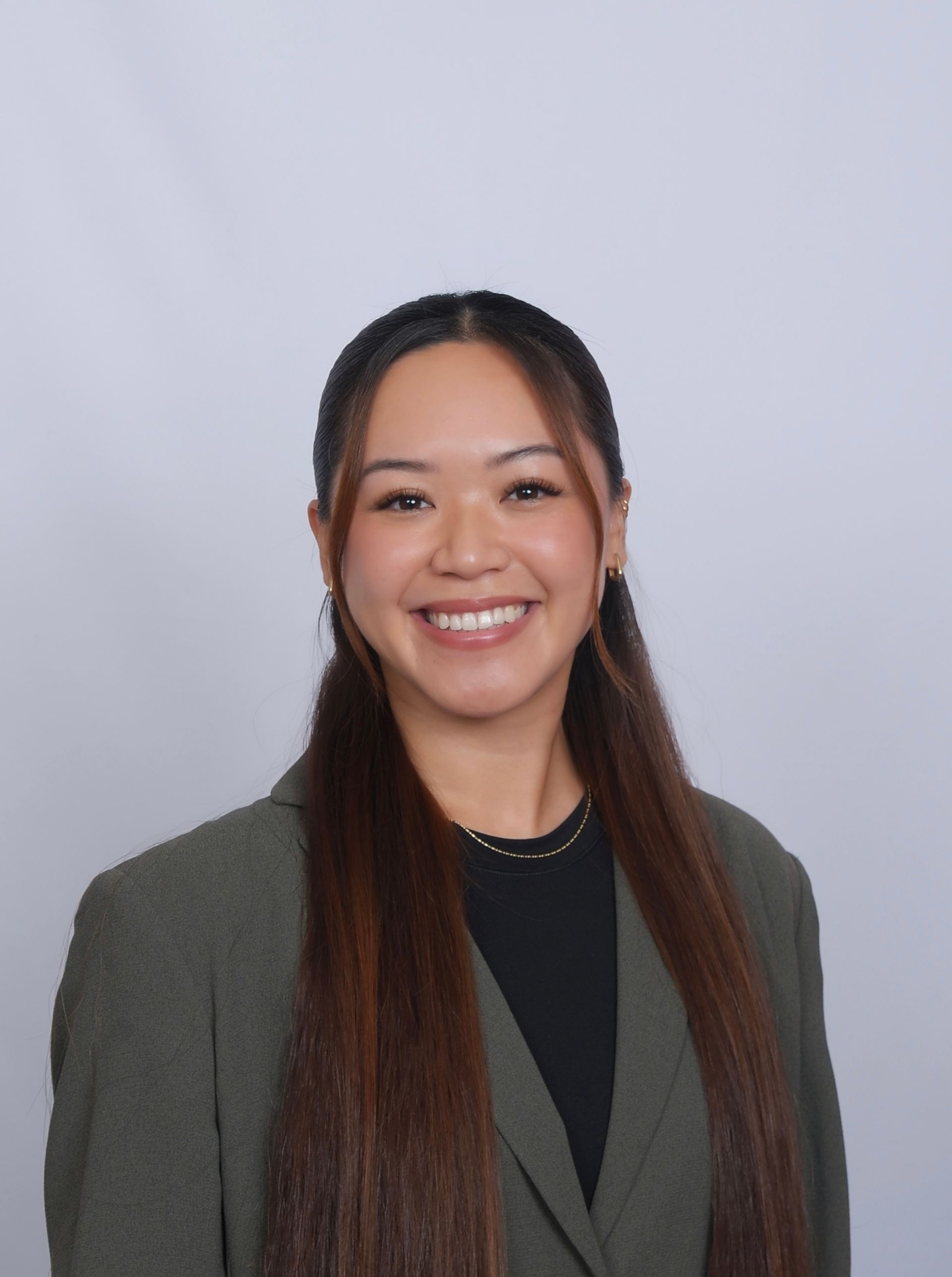 Woman with long brown hair, wearing a dark top and olive blazer, smiles at the camera.