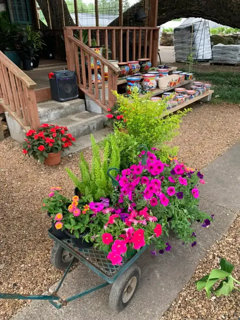A garden cart overflowing with colorful flowers sits outside a shop with steps and a wooden railing.