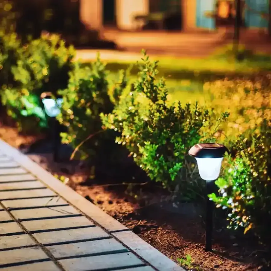 Solar lights illuminate a row of shrubs beside a brick walkway at dusk.