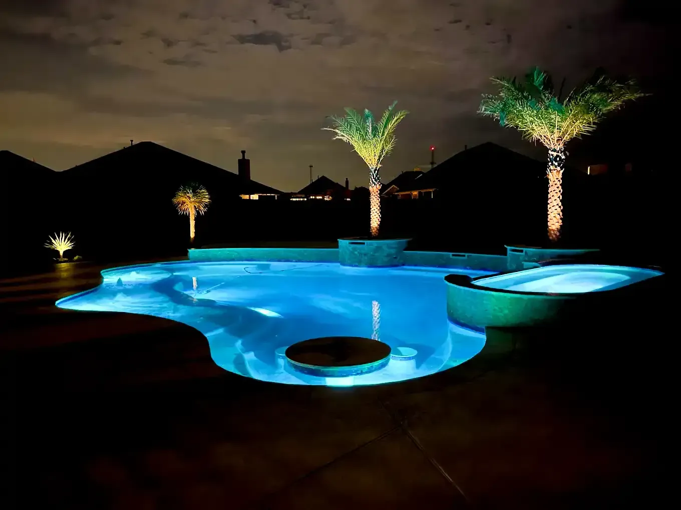 Nighttime shot of a lit-up swimming pool and hot tub, with palm trees and houses in the background.
